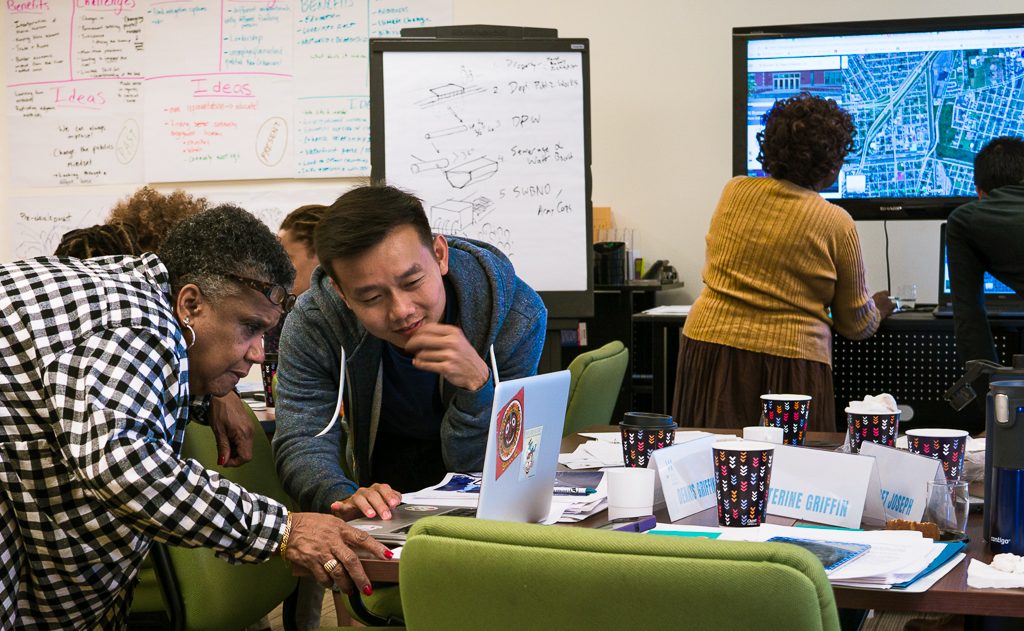 People collaborate in a planning workshop, with one group reviewing a laptop and another analyzing a large map on a screen, surrounded by notes, charts, and brainstorming materials on the walls.