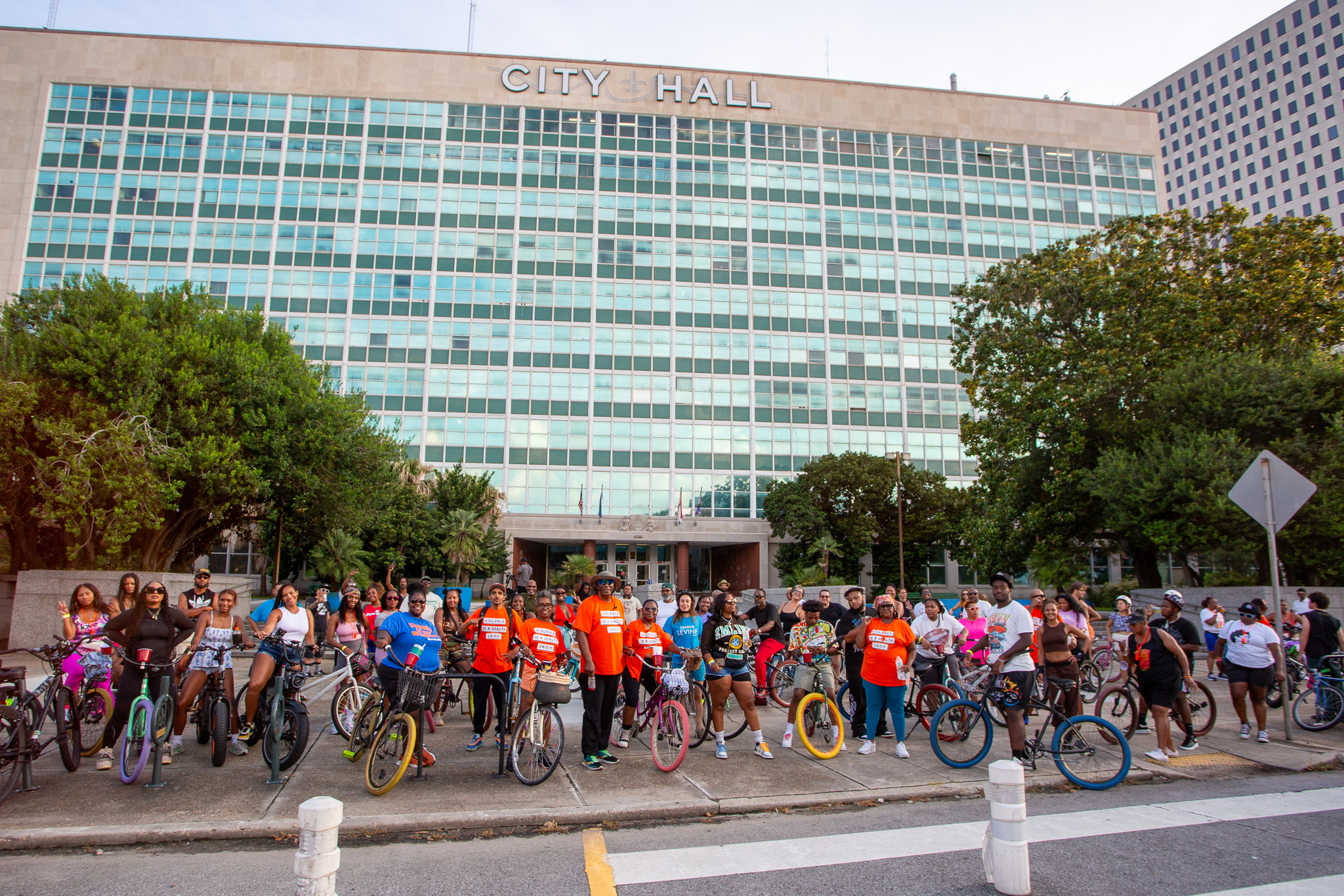 A large group of people stand with their bicycles in front of New Orleans City Hall during the Ride for Peace event. The diverse crowd includes men, women, and children wearing casual and event-themed clothing, some in orange shirts with slogans. The atmosphere is energetic and united, with participants posing for a photo before the community bike ride begins. The tall, glass-paneled City Hall building looms in the background, flanked by large trees.