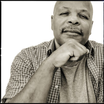 Portrait of Calvin Duncan, author of "The Jailhouse Lawyer," gazing confidently at the camera with his hand resting under his chin. He wears a checkered shirt over a t-shirt, and the background is a clean, bright white, emphasizing his thoughtful expression.